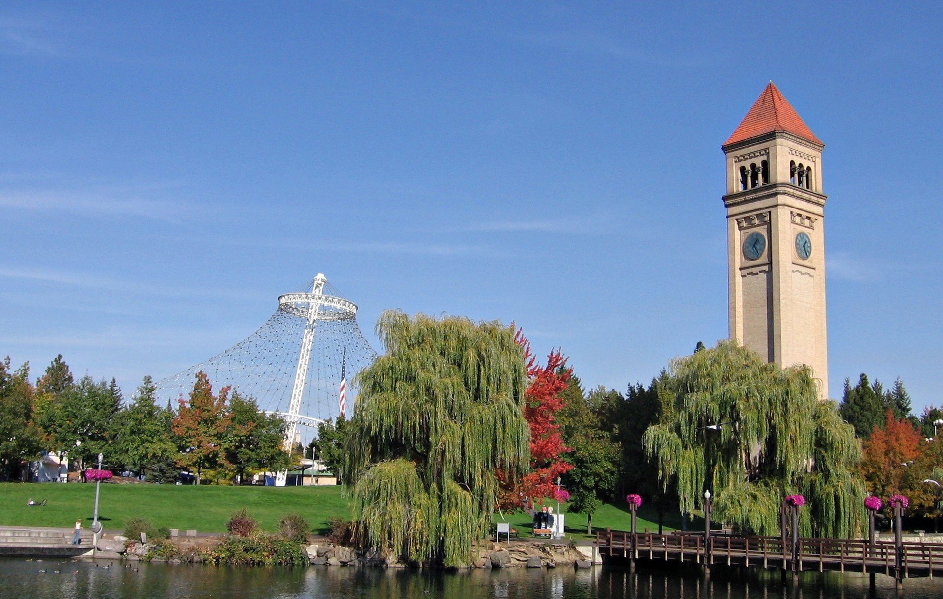 Riverfront park with white illuminated cross, historic clock tower with red roof, green trees, and autumn foliage under blue sky